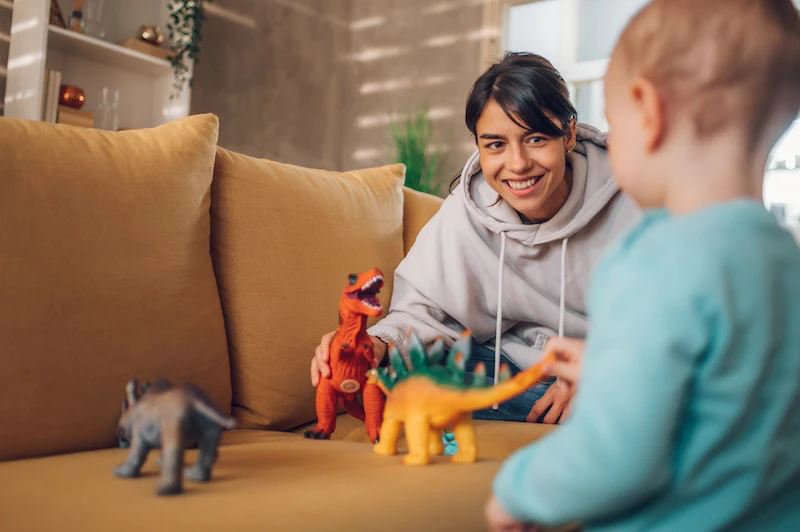 A young girl playing with a toddler in a living room with toy dinosaurs.