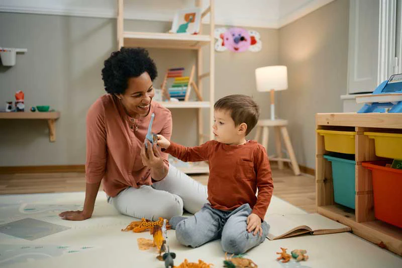 Childcarer playing with a young boy on the floor in a child's playroom.