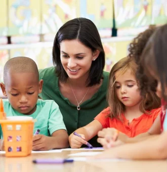 Young woman teaching and coloring with young kids in a classroom setting.