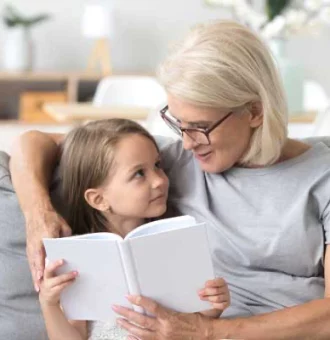 Loving grandmother teaching granddaughter reading book sitting on sofa.